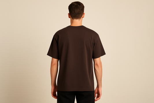 Professional e-commerce product photography of a male model wearing an oversized dark brown crew neck t-shirt, back view with model standing straight facing away from camera with hands relaxed at sides, wearing dark pants against a neutral beige background with studio lighting