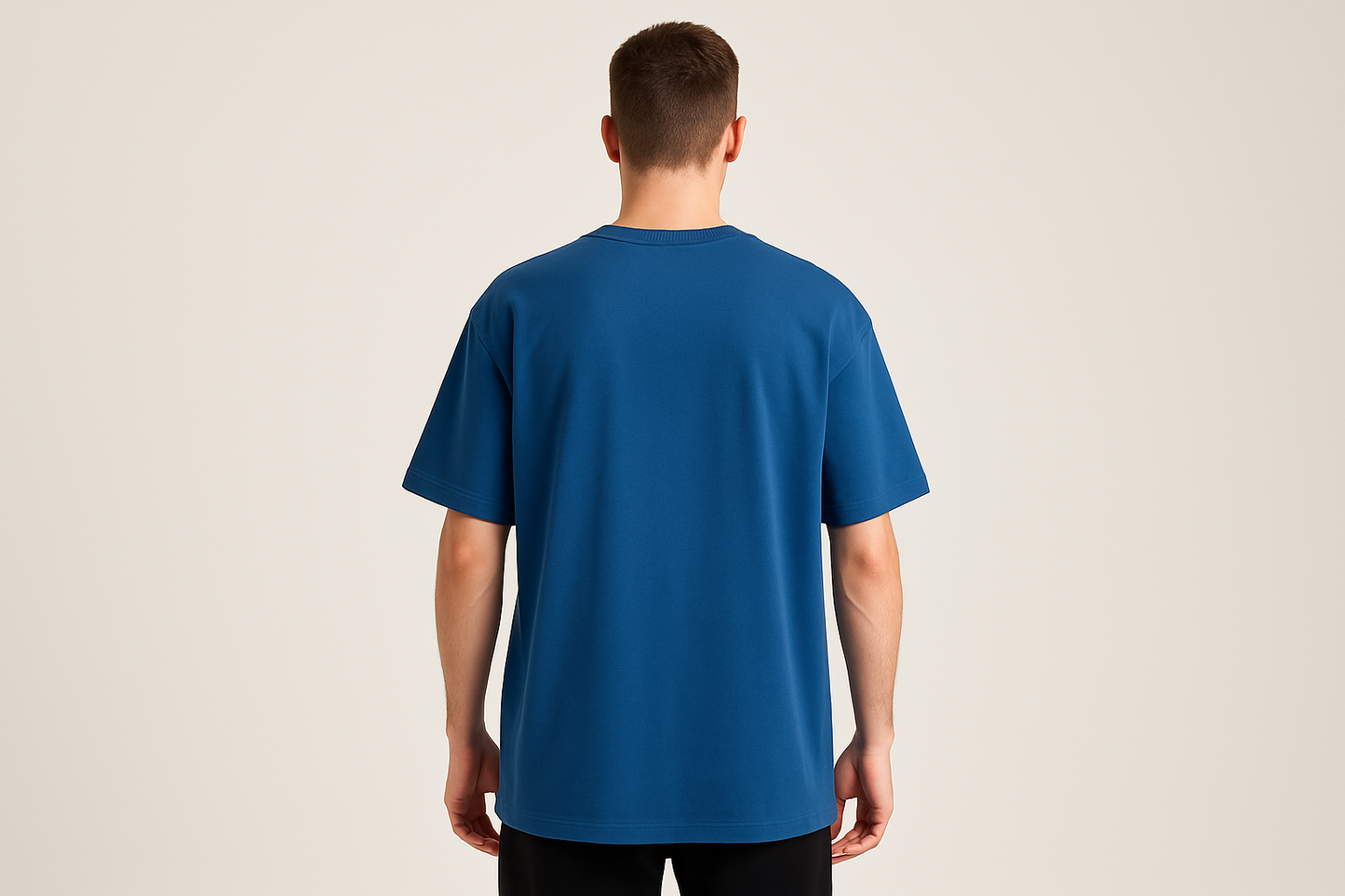 Professional e-commerce product photography of a male model wearing an oversized royal blue crew neck t-shirt, back view with model standing straight facing away from camera with hands relaxed at sides, wearing dark pants against a neutral beige background with studio lighting