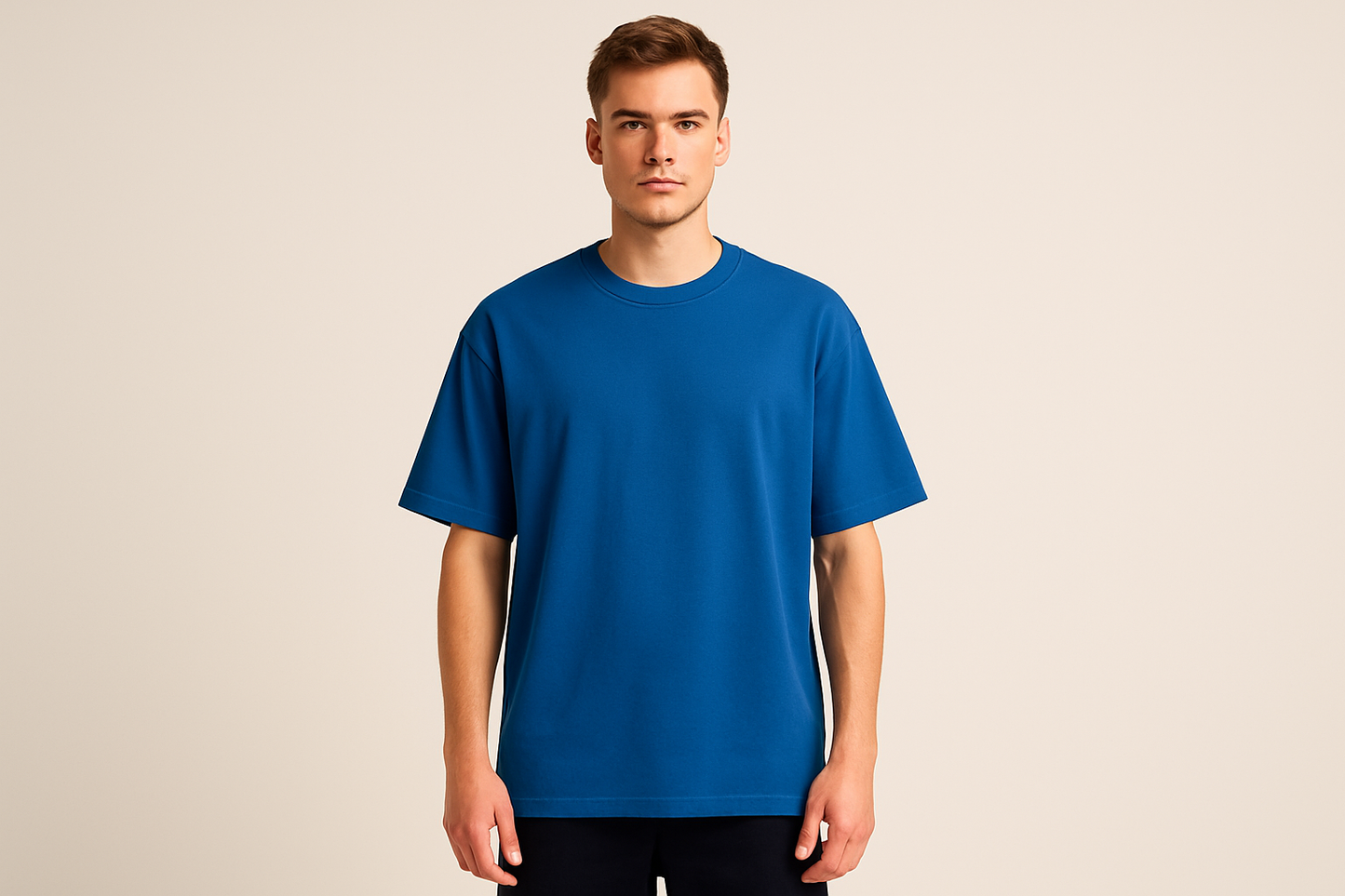 Professional e-commerce product photography of a male model wearing an oversized royal blue crew neck t-shirt, standing straight facing camera with hands relaxed at sides, wearing dark pants against a neutral beige background with studio lighting