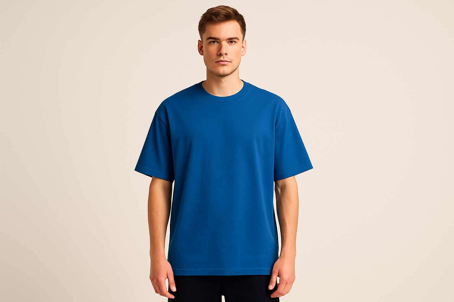 Professional e-commerce product photography of a male model wearing an oversized royal blue crew neck t-shirt, standing straight facing camera with hands relaxed at sides, wearing dark pants against a neutral beige background with studio lighting
